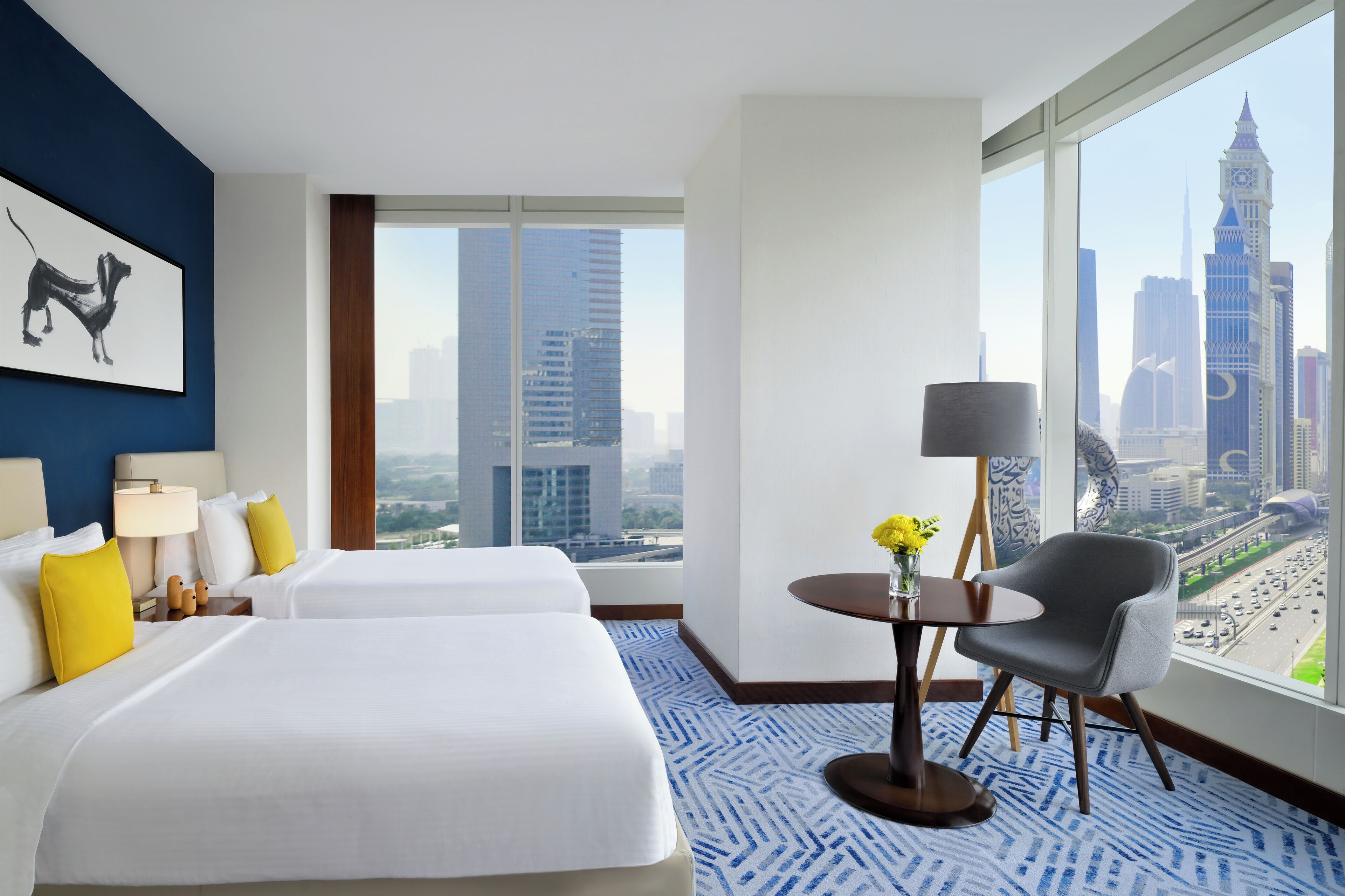 Une chambre d’hôtel avec deux lits, une table avec une chaise, une lampe et une vue sur le paysage urbain.