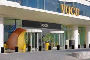 A modern hotel entrance with a prominent 'VOCO' sign, glass doors, and a unique yellow and brown sculpture.