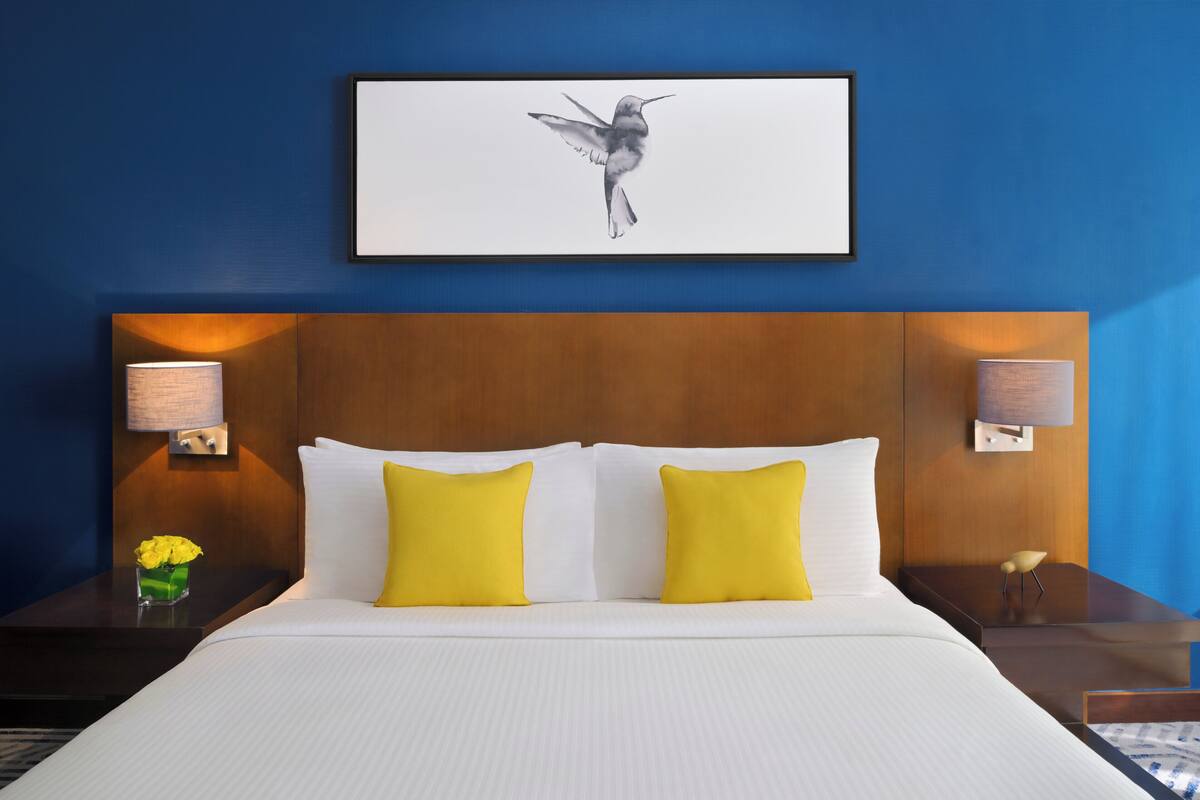 A hotel room with a large bed, two yellow pillows, a wooden headboard, and a framed artwork of a hummingbird on the wall.