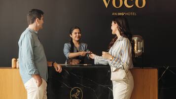 Mostrador de recepción con un logotipo, un hombre y una mujer de pie, y un cartel con el nombre del hotel.