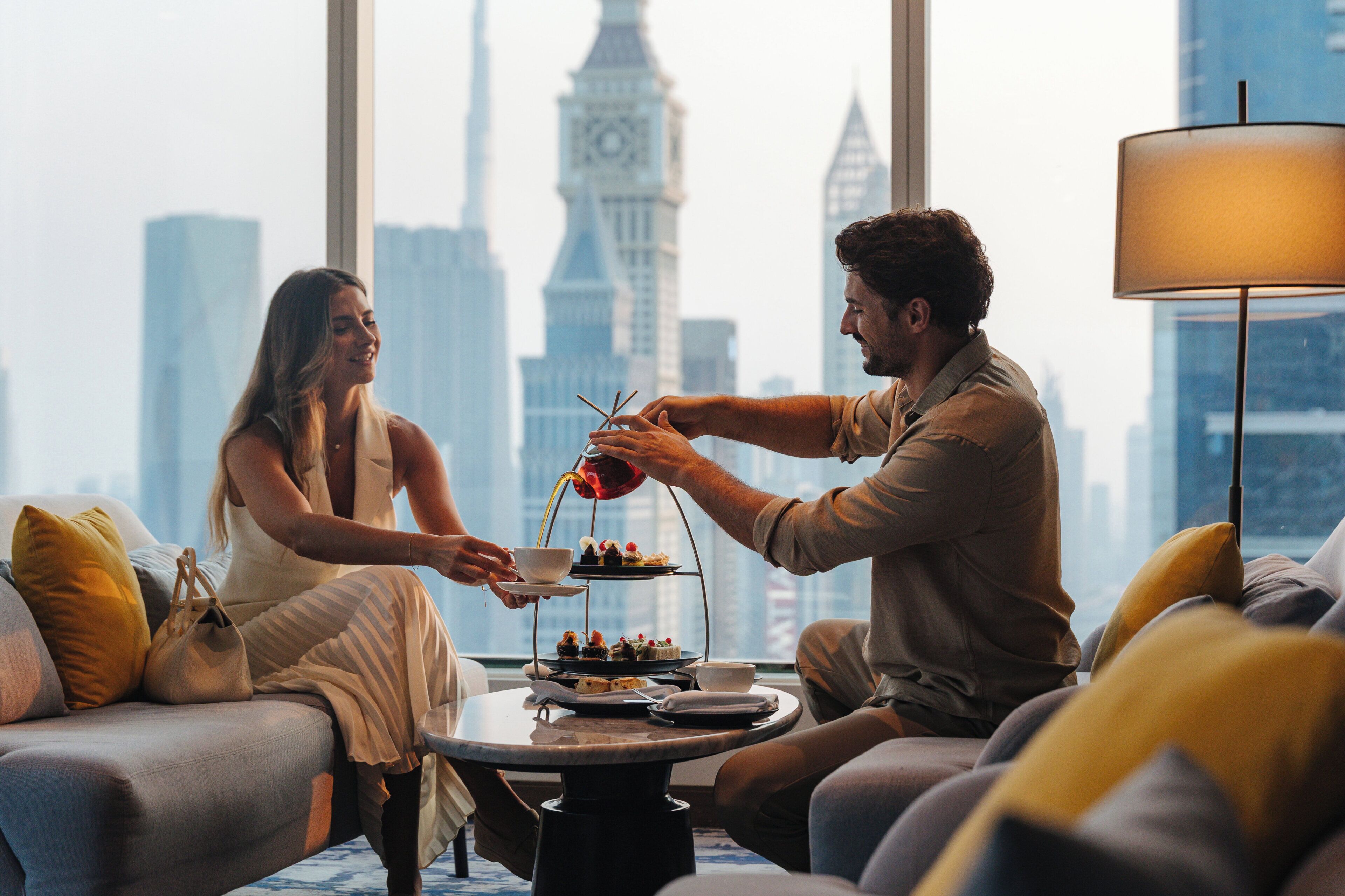 A couple seated on a sofa with a city skyline view, a table with a tea set, and a lamp.