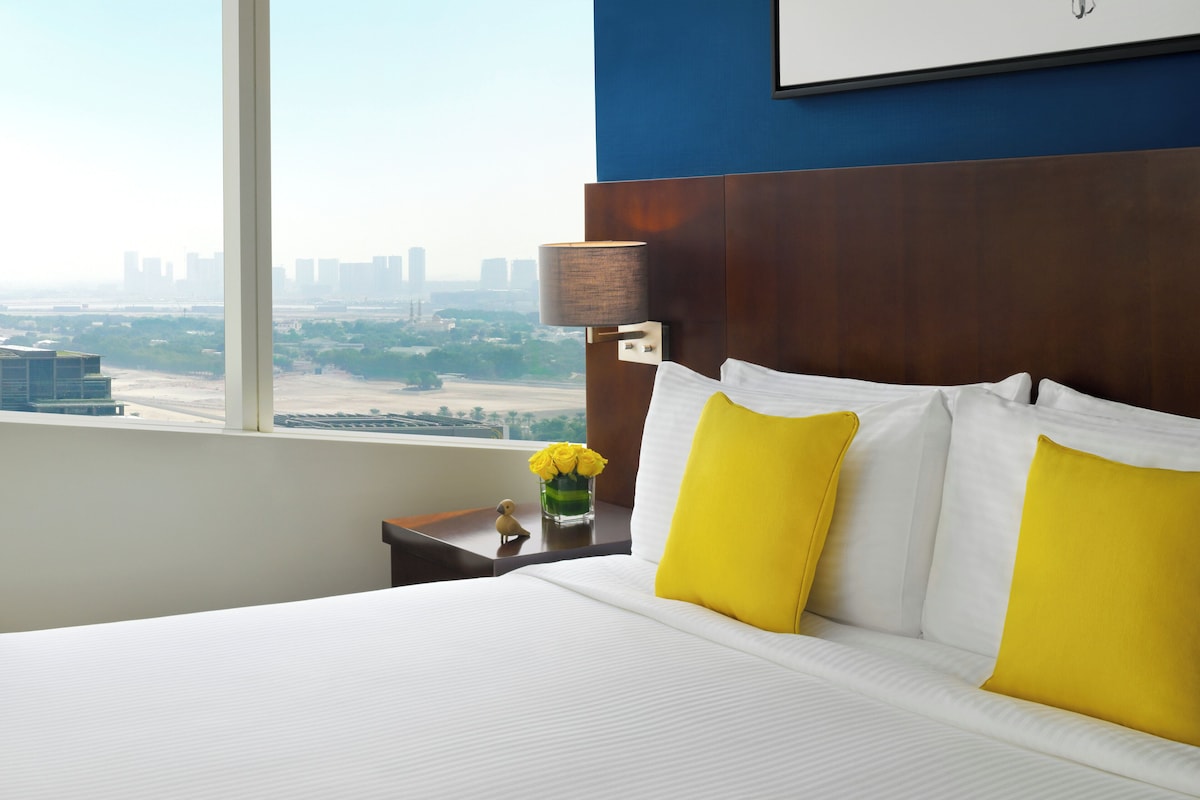 A hotel room with a large bed, a nightstand with a lamp, and a view of a cityscape through a large window.