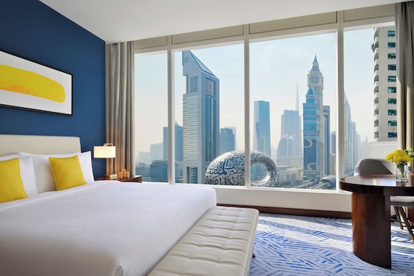 A hotel room with a large bed, a view of a city skyline through floor-to-ceiling windows, a round table with a vase of flowers, and a blue patterned rug.