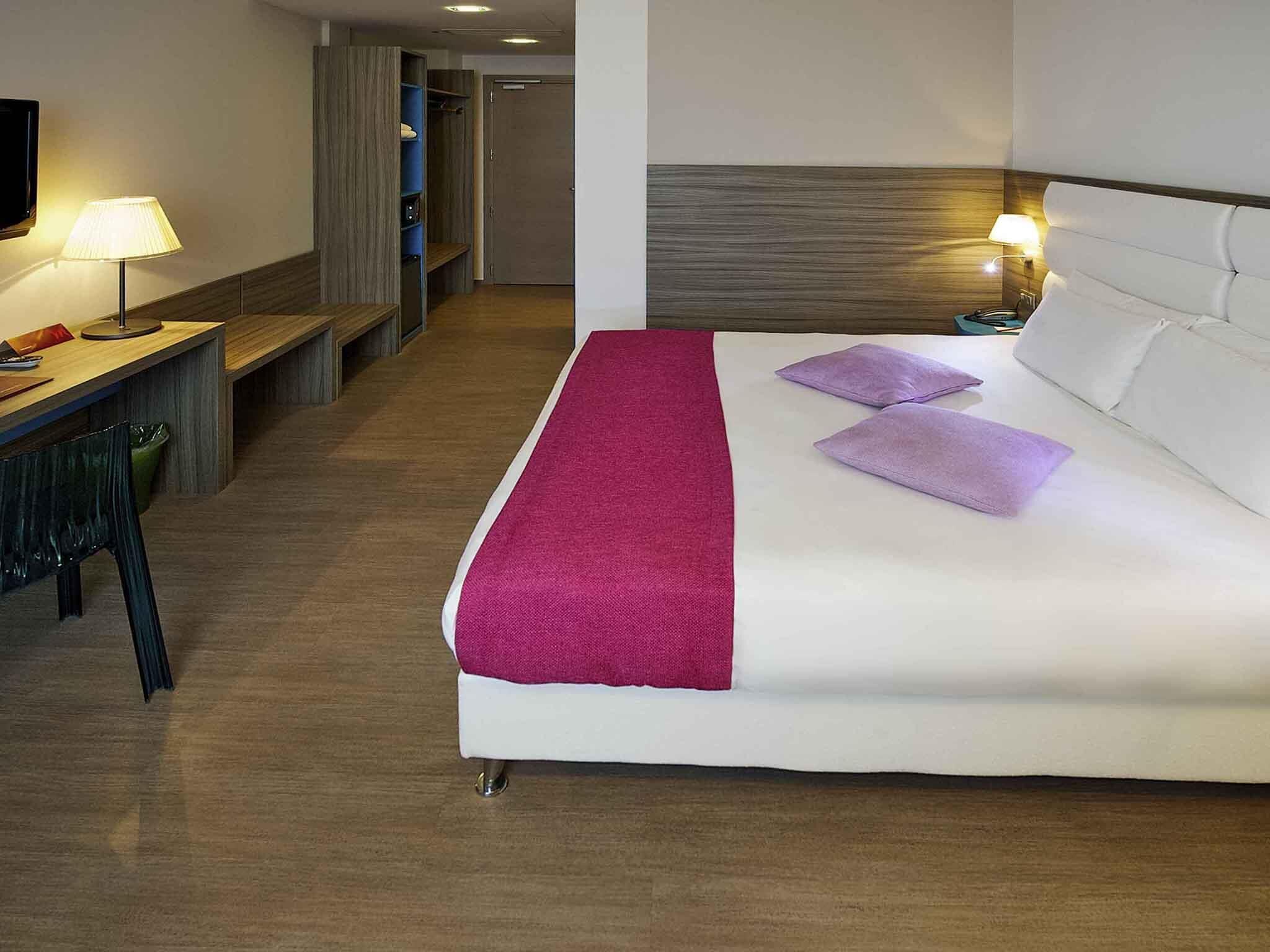 Una camera d'albergo con un letto, un comodino, una lampada e una scrivania con una sedia.