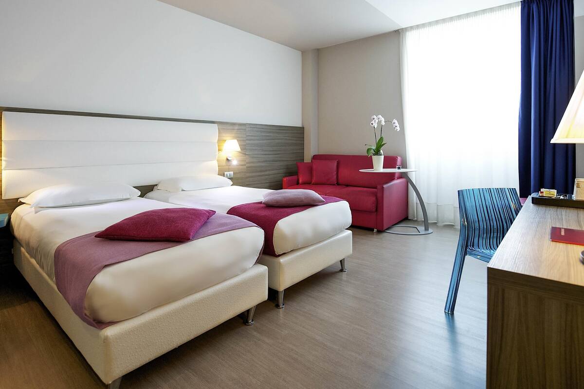 Een hotelkamer met een groot bed, een rode bank, een tafeltje met een lamp en een stoel.