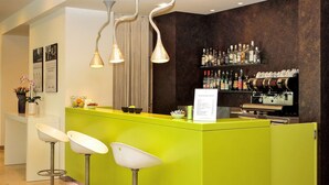 Un espace bar avec un comptoir vert, trois tabourets de bar blancs, un panneau affichant le menu et un mur recouvert de bouteilles d’alcool.