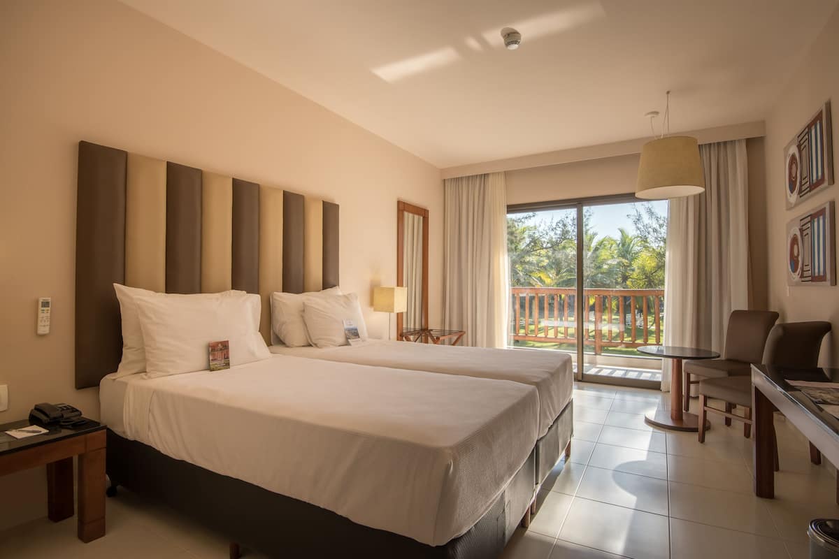 Hotelkamer met twee bedden, een bureau, een stoel, een balkon met uitzicht op groen en een aan het plafond bevestigde beveiligingscamera.