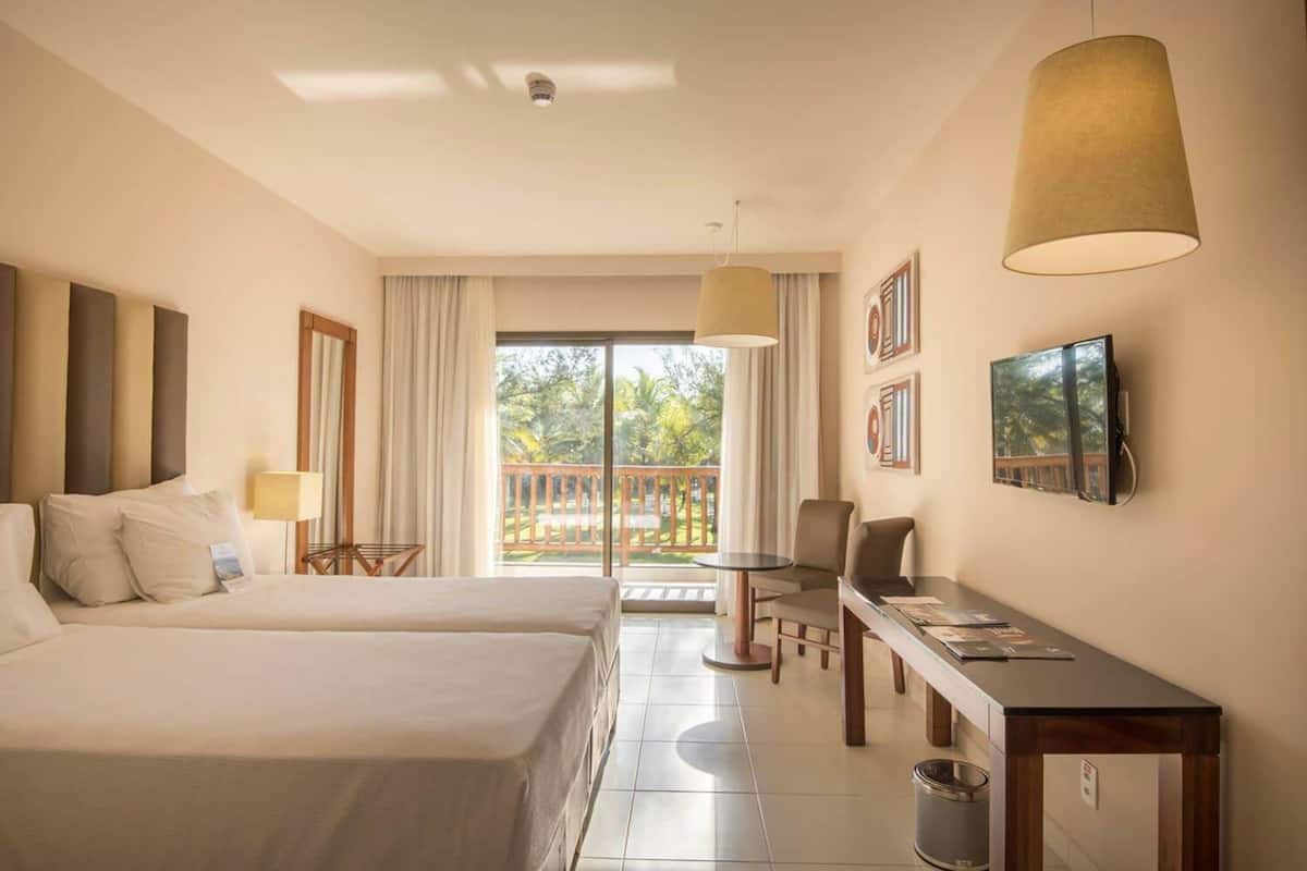 Hotelkamer met twee bedden, een bureau, een stoel, een televisie en een balkon met uitzicht op groen.