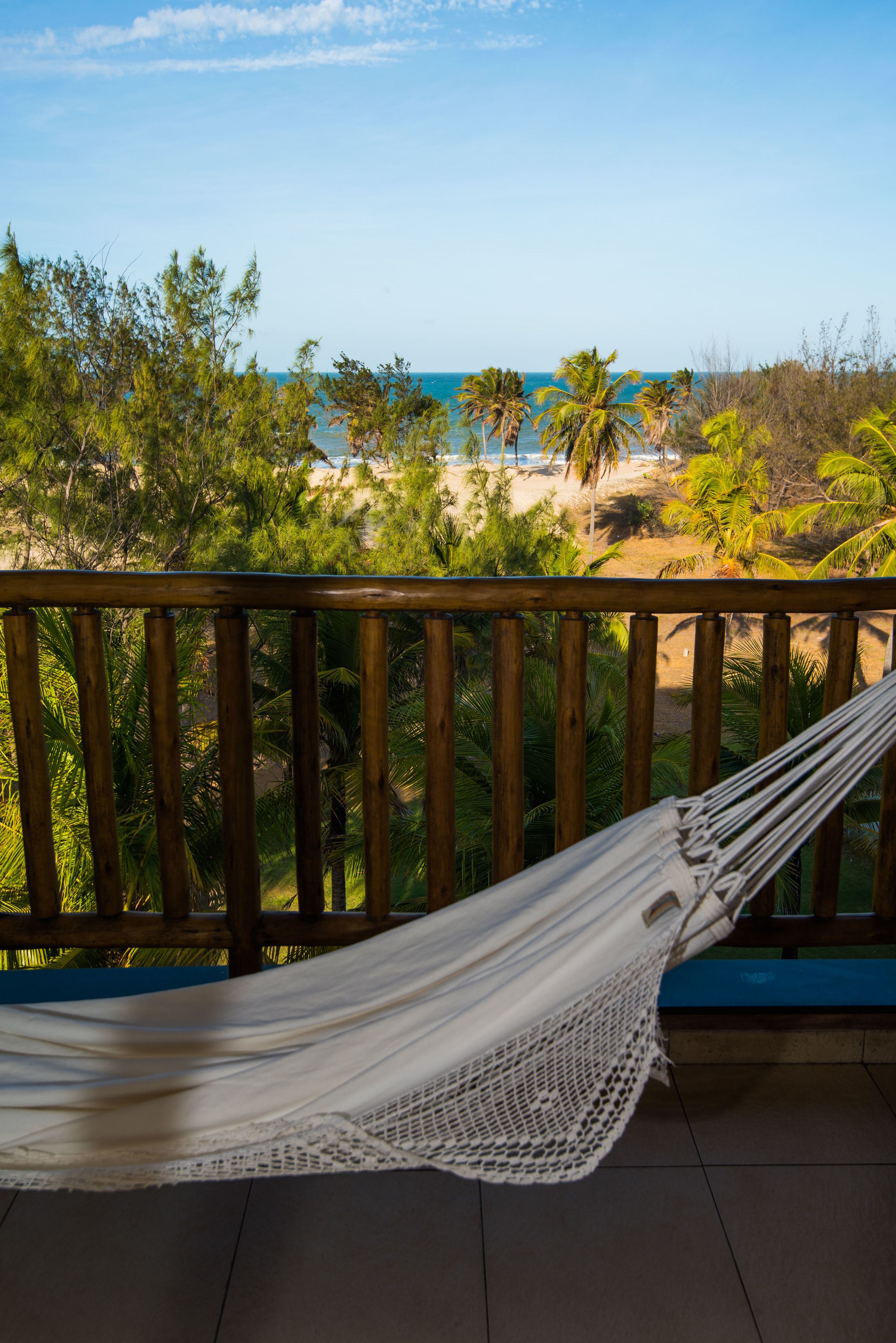 A hammock on a wooden deck overlooking a beach with palm trees and the ocean.