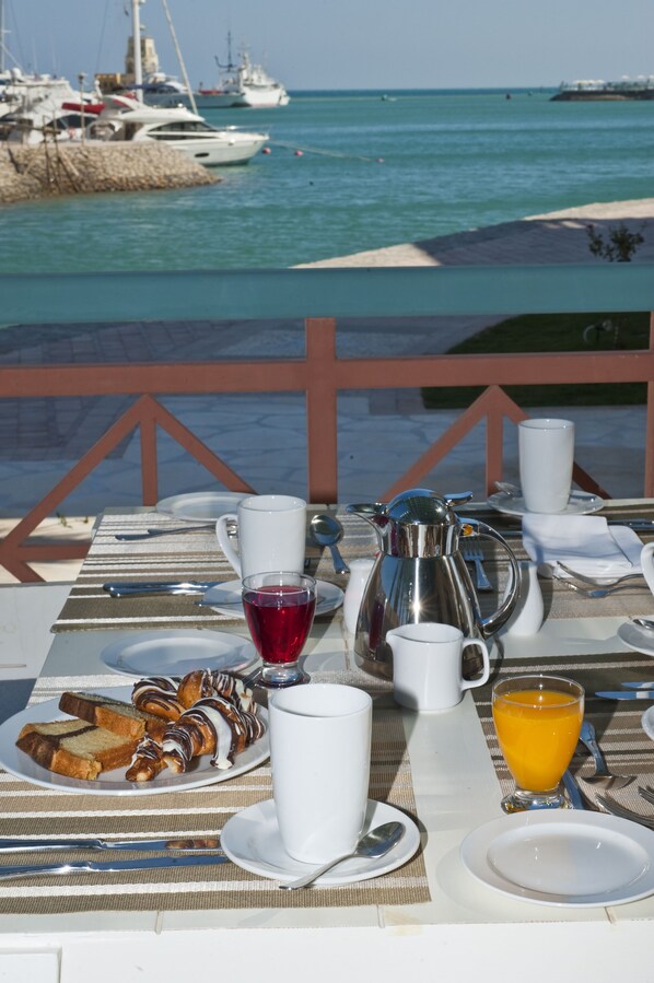 Ein Frühstückstisch mit Blick auf das Meer und Boote, mit einer weißen Kaffeekanne, einem Glas Orangensaft und einem Teller Gebäck.