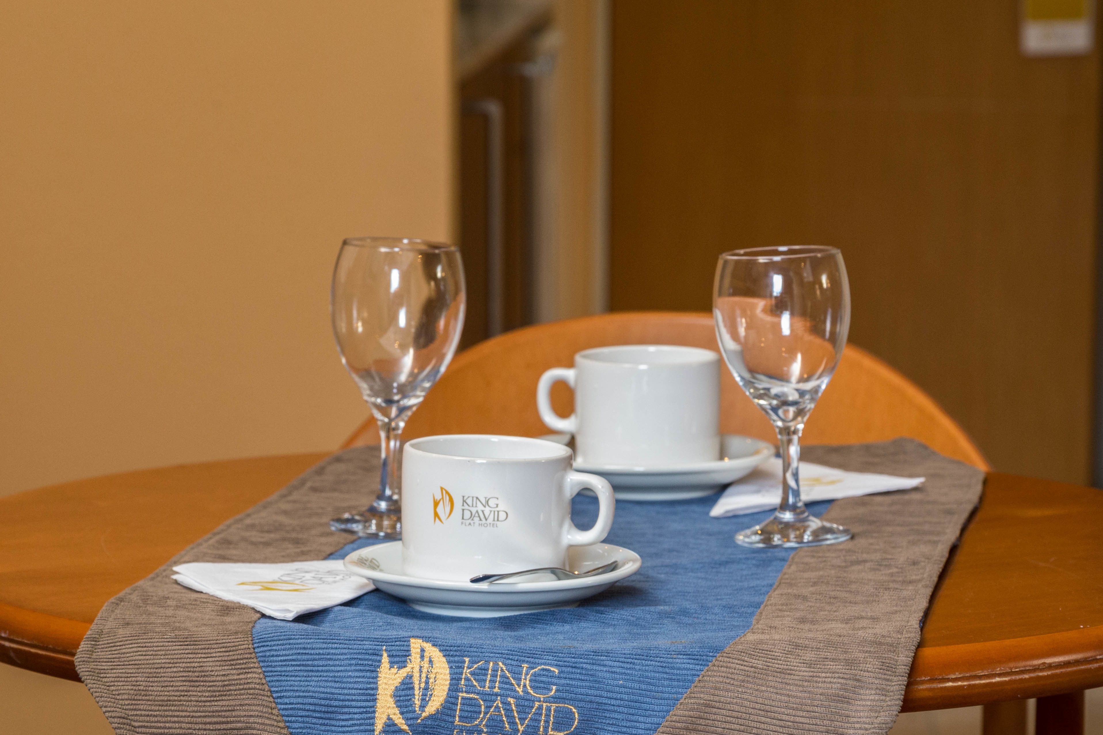 A dining table with two white cups and two wine glasses, a napkin, and a placemat with the King David logo.