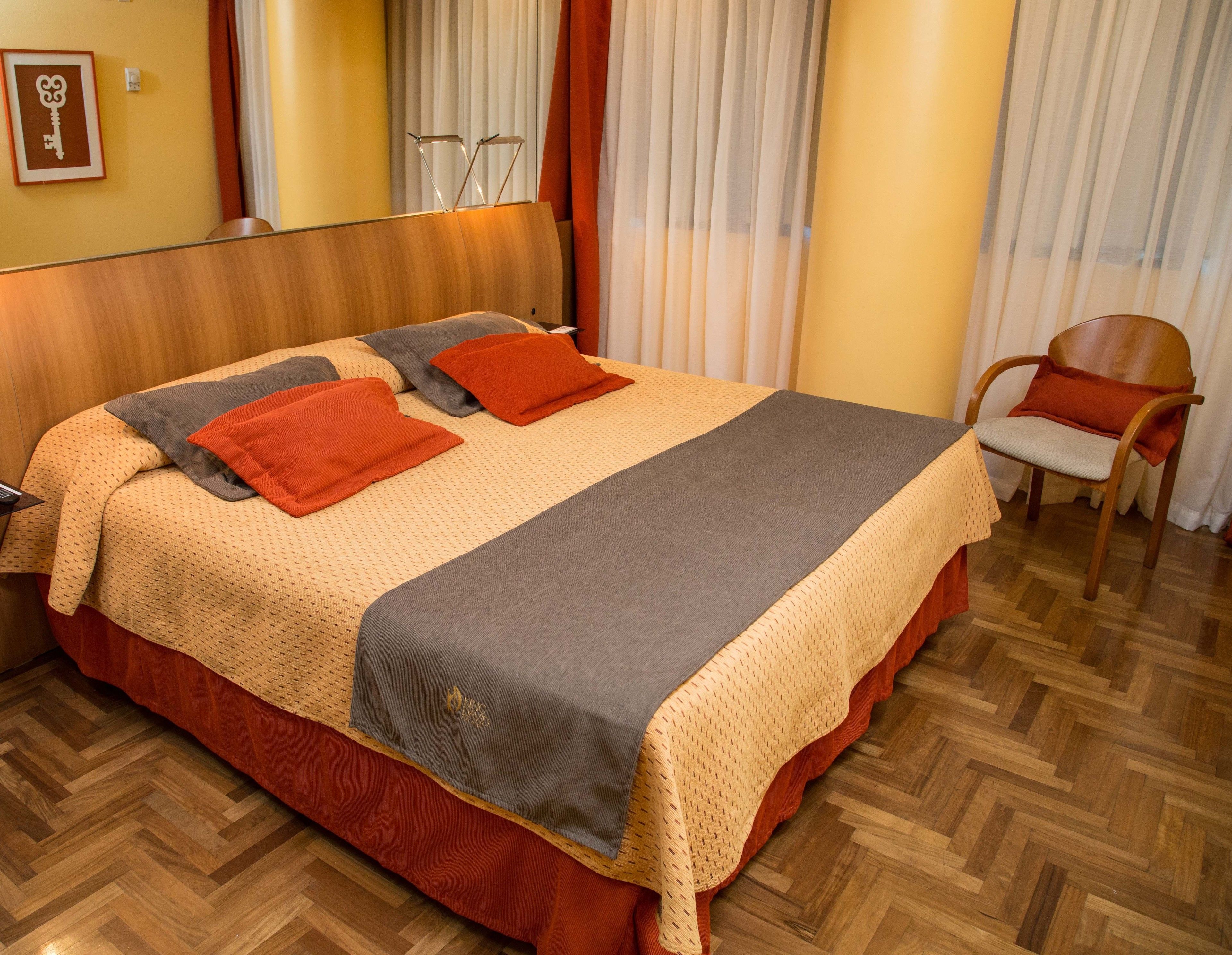 Un letto ben rifatto con una coperta a fantasia e due cuscini, una testiera in legno, una sedia con un cuscino e un pavimento in legno.