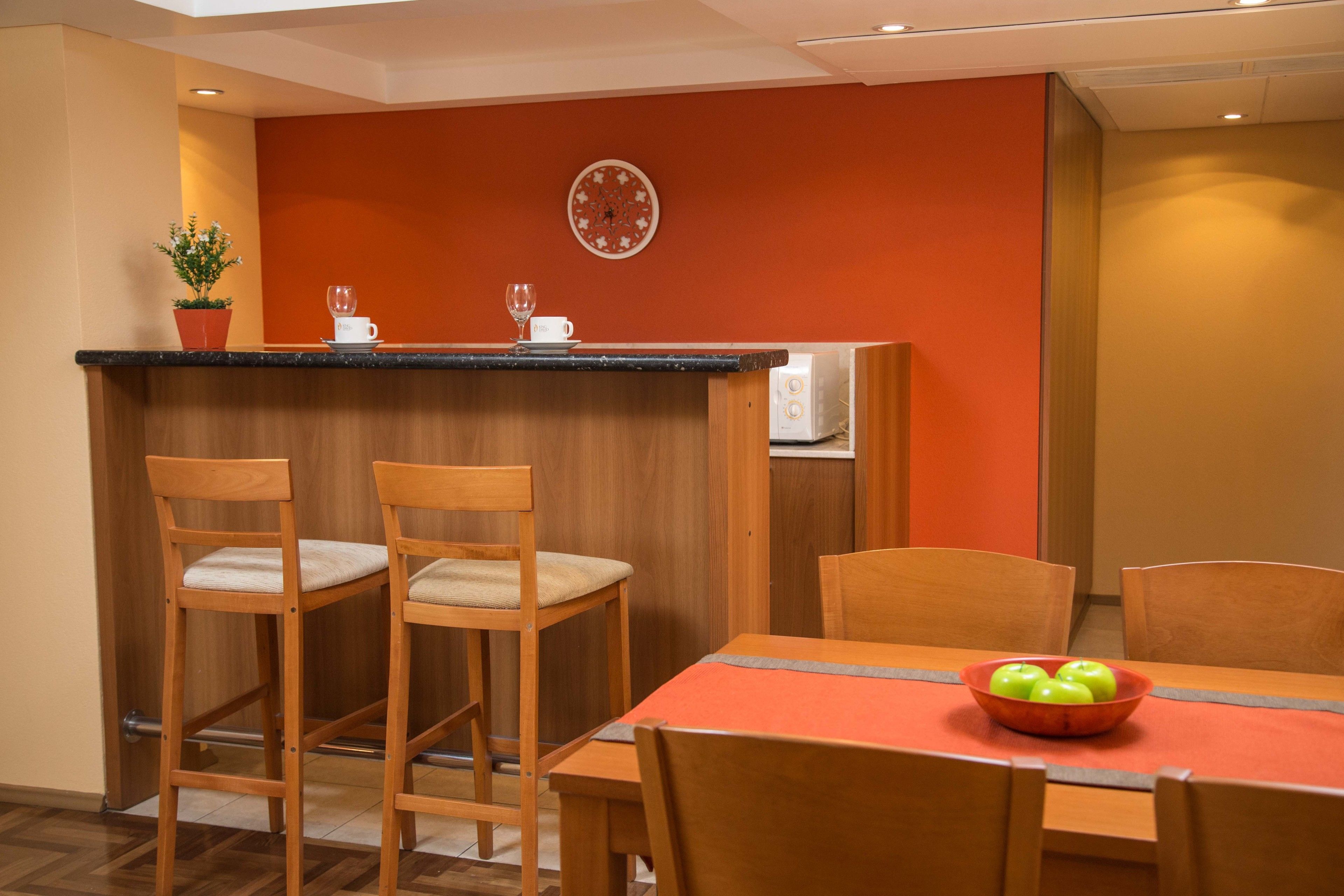 A dining area with a wooden table and chairs, a bowl of green apples, a potted plant, and a microwave in the background.