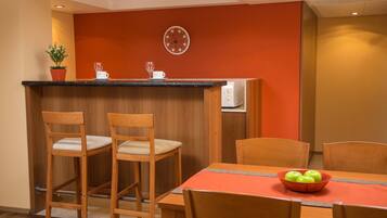 A dining area with a wooden table and chairs, a bowl of green apples, a potted plant, and a microwave in the background.