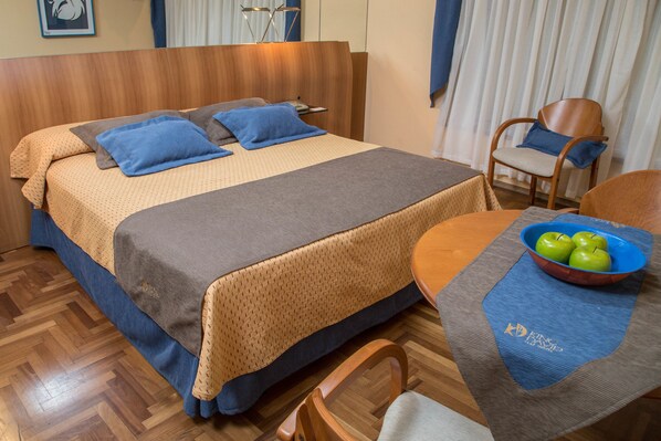 A hotel room with a wooden headboard, a bed with blue and brown bedding, a round table with a bowl of green apples, and a chair. - King David Flat Hotel (Córdoba)