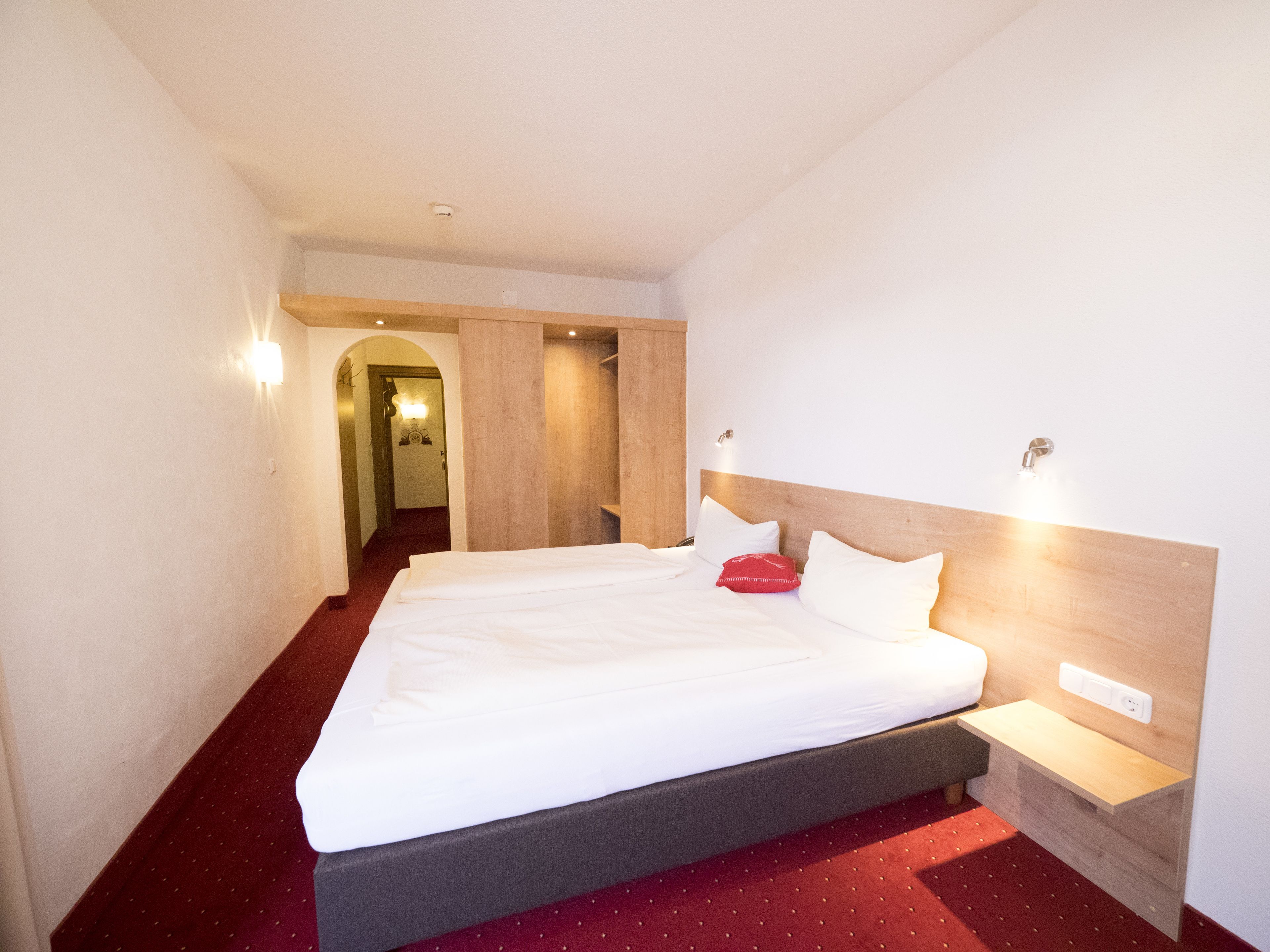A hotel room with a large bed, a wooden headboard, a bedside table, and a red carpet.