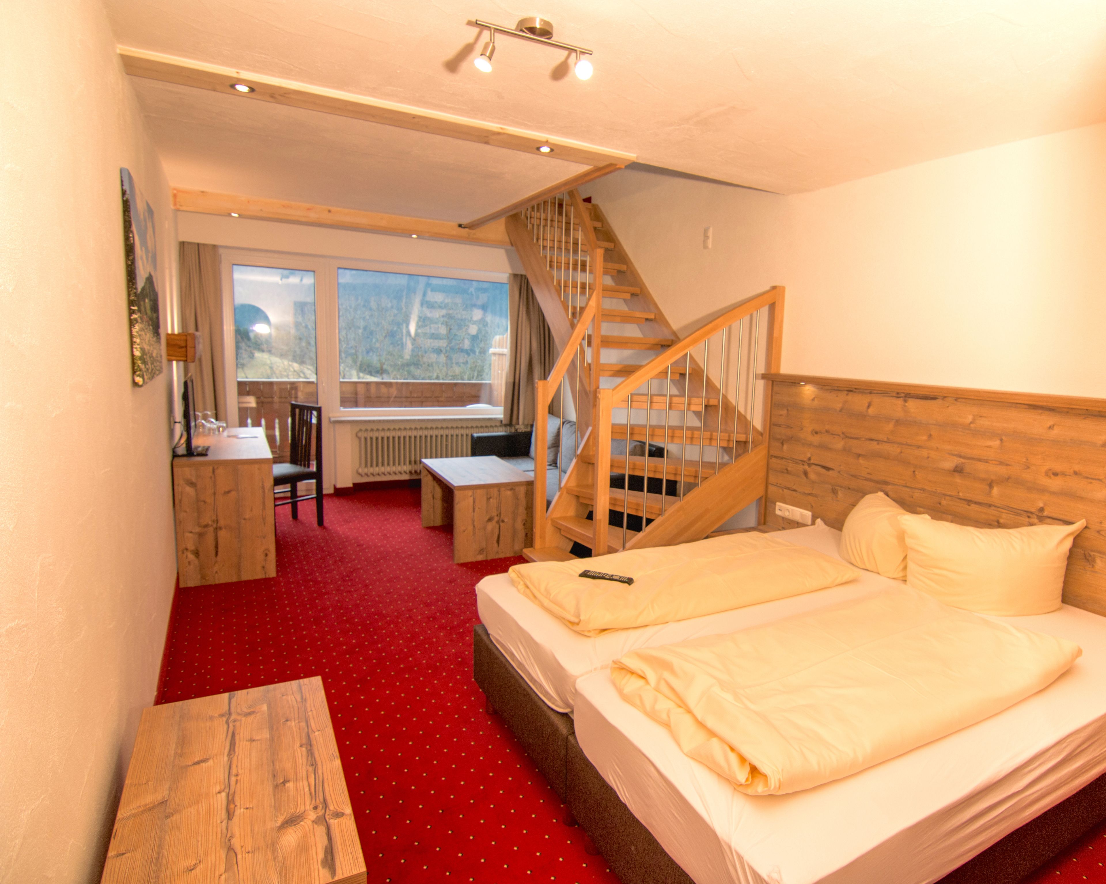 A bedroom with a bed, a desk, a chair, a wooden table, a staircase, and a window with a view.