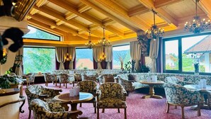 A spacious dining area with wooden ceiling beams, floral-patterned chairs, round tables, and large windows offering a view of the outdoors. - Berghotel Tirol (Jungholz)