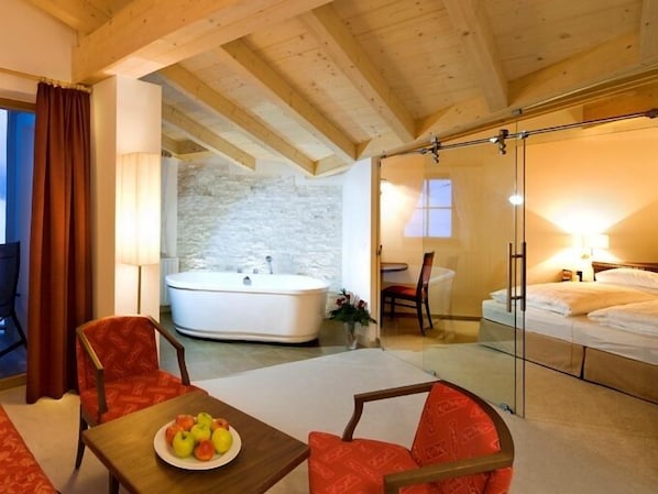 A hotel room with a large bathtub, a separate shower area, a dining table with fruit, and a bed.