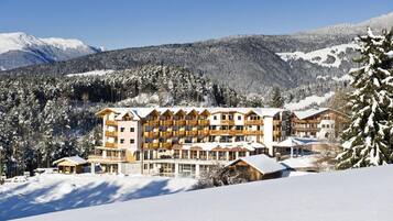 A multi-story hotel with a traditional Alpine architectural style, featuring balconies and large windows, set against a snowy mountainous backdrop.