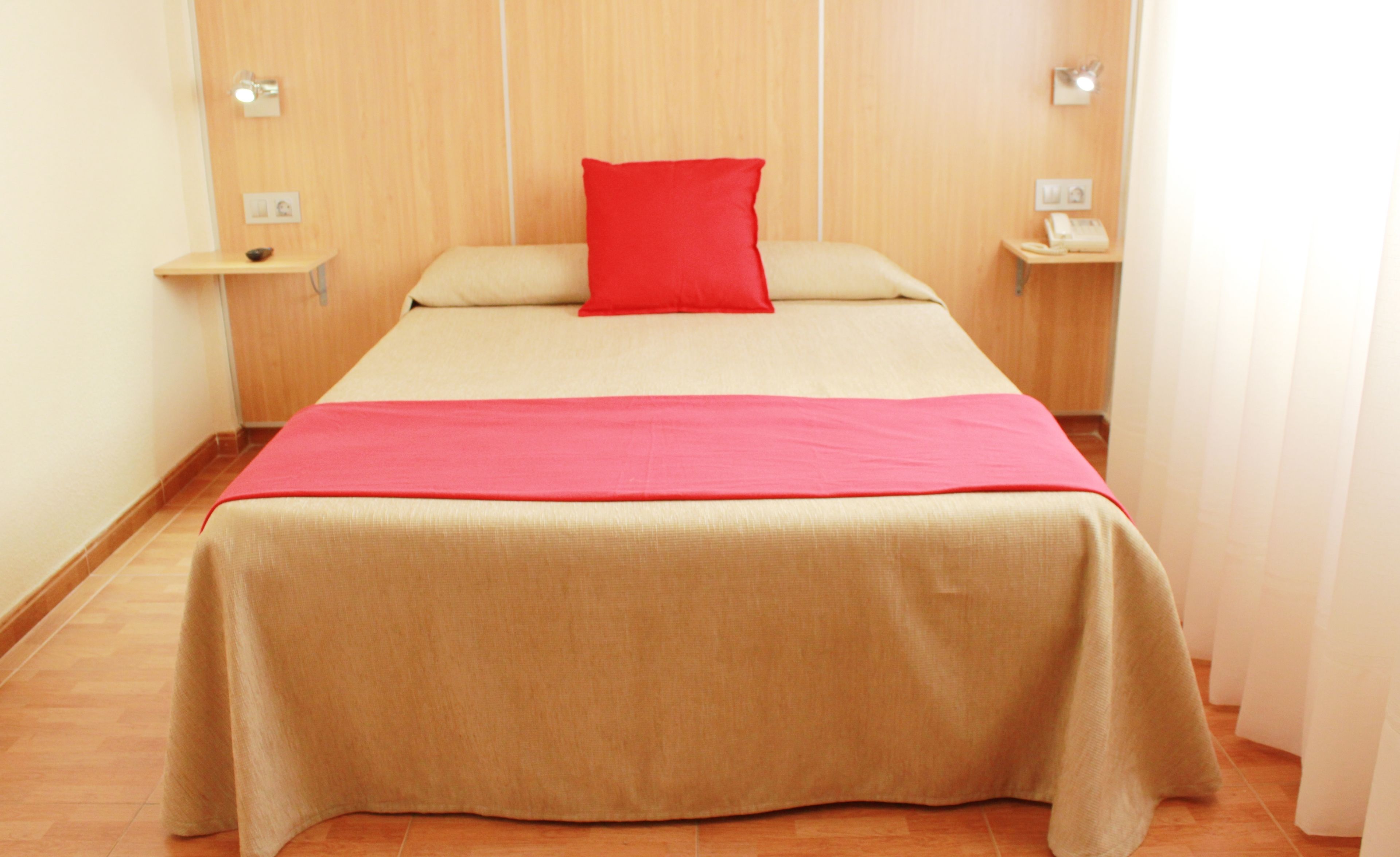 A neatly made bed with a red pillow and a beige bedspread in a room with wooden walls and a wooden floor.