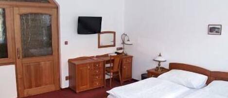 A bedroom with a wooden wardrobe, a desk, a chair, a bed, a television, and a small table with a lamp.