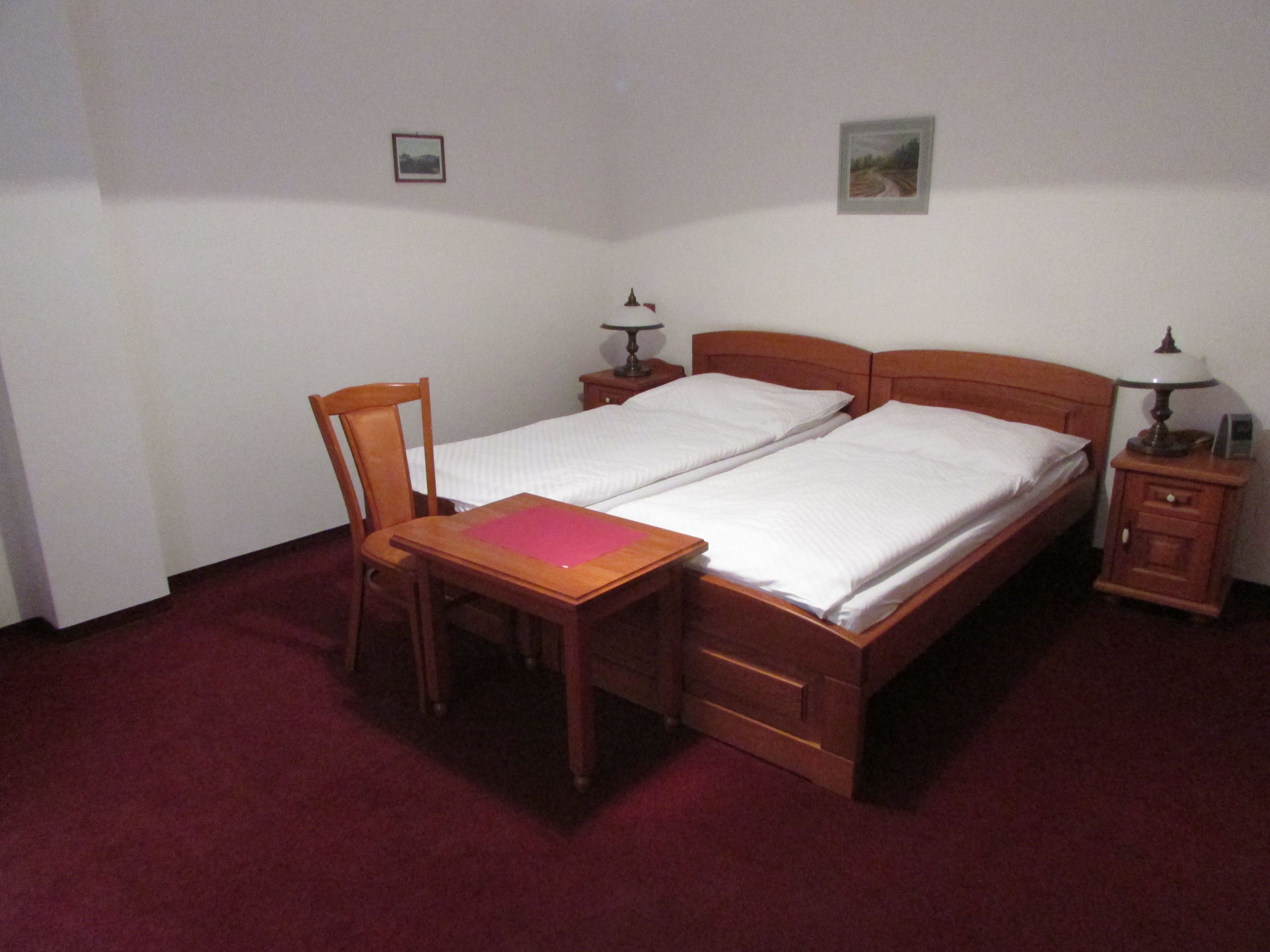A double bed with a wooden headboard, a small bedside table with a lamp, a chair, and a desk with a pink folder.