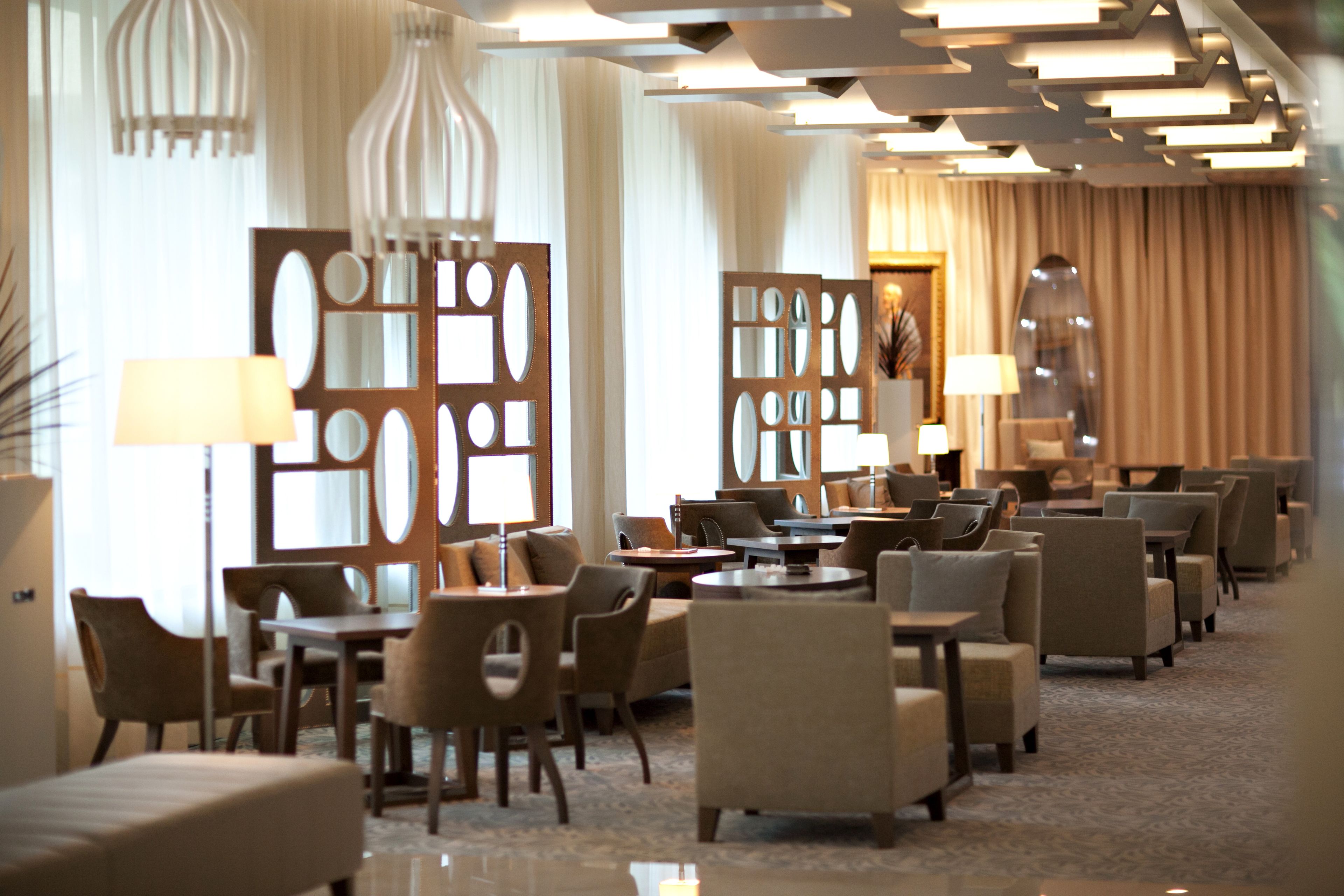 A hotel lobby with multiple seating areas, including sofas and armchairs, arranged around tables. There are decorative panels with cut-out designs and a large mirror on the wall.