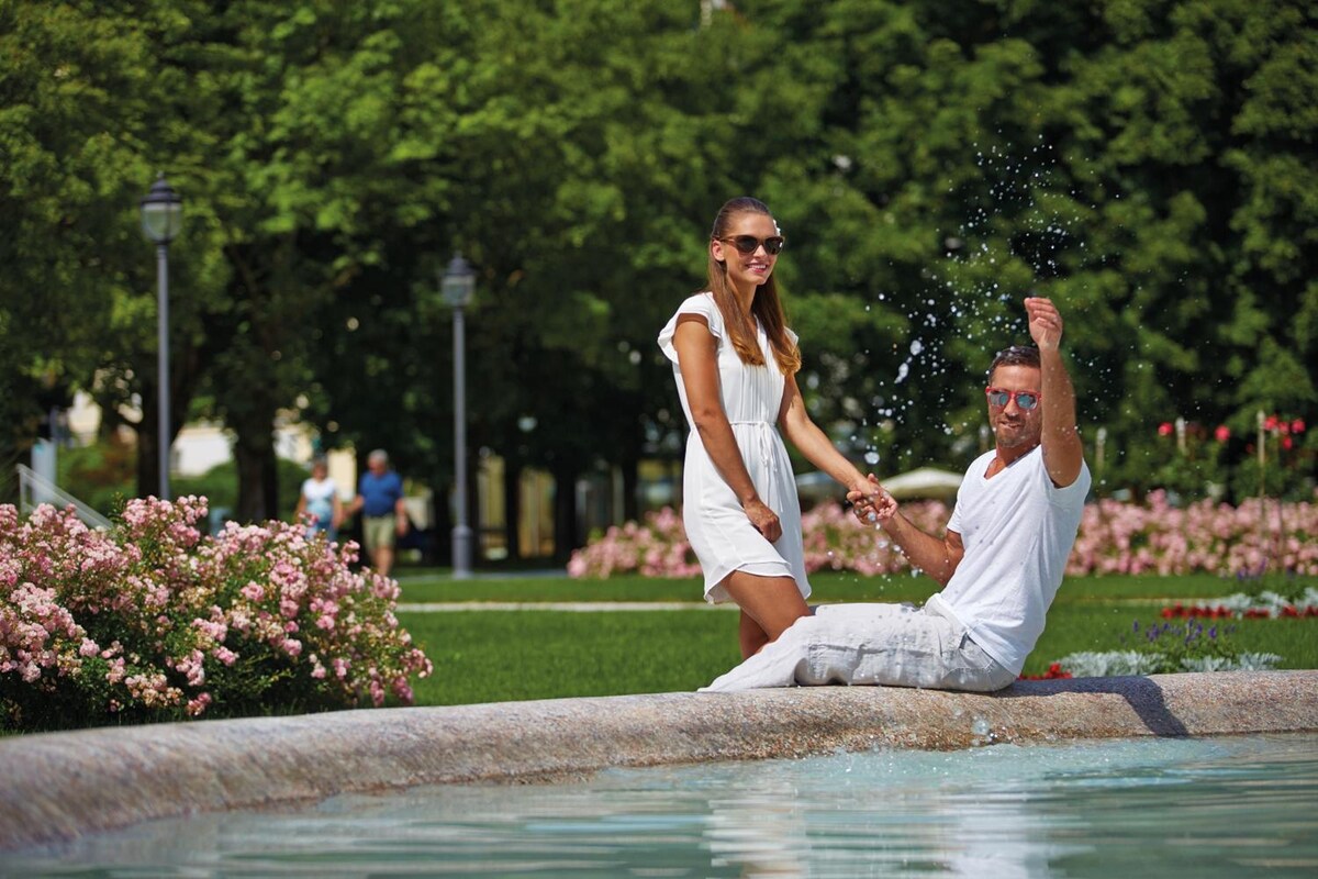 Una pareja junto a una fuente en un parque con follaje y flores.