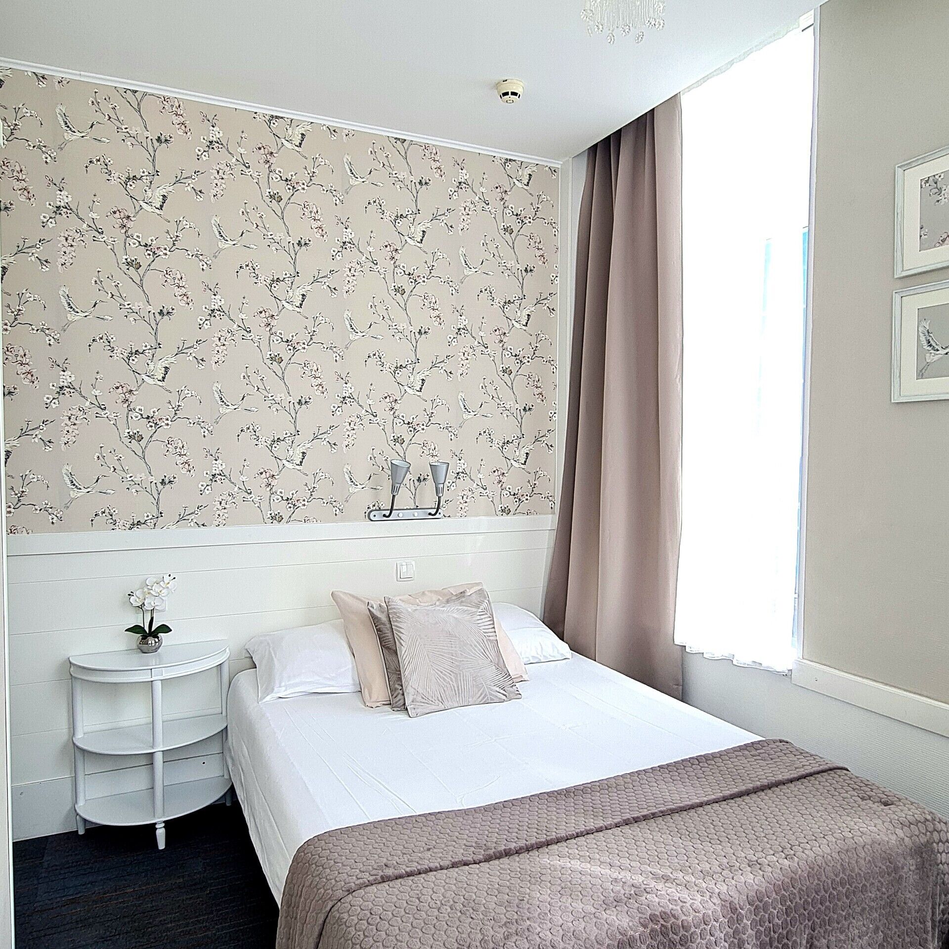 Ein Hotelzimmer mit floralem Tapetenmuster, einem Bett mit weißer und beiger Bettwäsche, einem Nachttisch mit Blumenvase und einem Fenster mit Vorhängen.