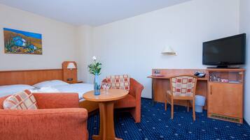 Habitación de hotel con cama, dos sillones, una mesita con un florero, un escritorio con una silla, un televisor y un cuadro en la pared.