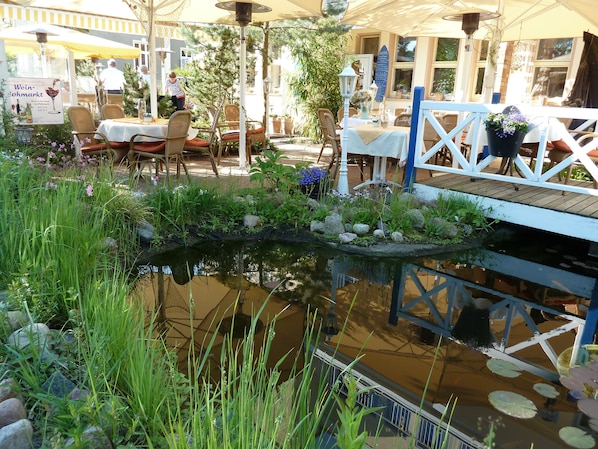 An outdoor seating area with tables and chairs, a small pond with rocks and plants, and a wooden deck with a railing. - Vineta Hotels (Zinnowitz)