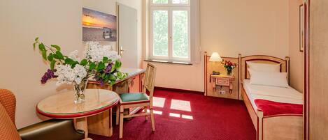 A hotel room with a bed, a desk with a chair, a small table with a vase of flowers, and a window with a view of the sunset.