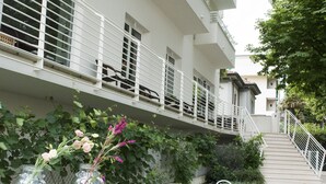 A white outdoor seating area with a table and chairs, a staircase leading to a balcony, and a lush green garden. - Hotel Houston (Rimini)
