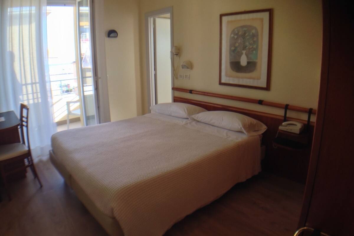 Una habitación de hotel con una cama grande, una silla, una mesita pequeña, un teléfono y un cuadro en la pared.