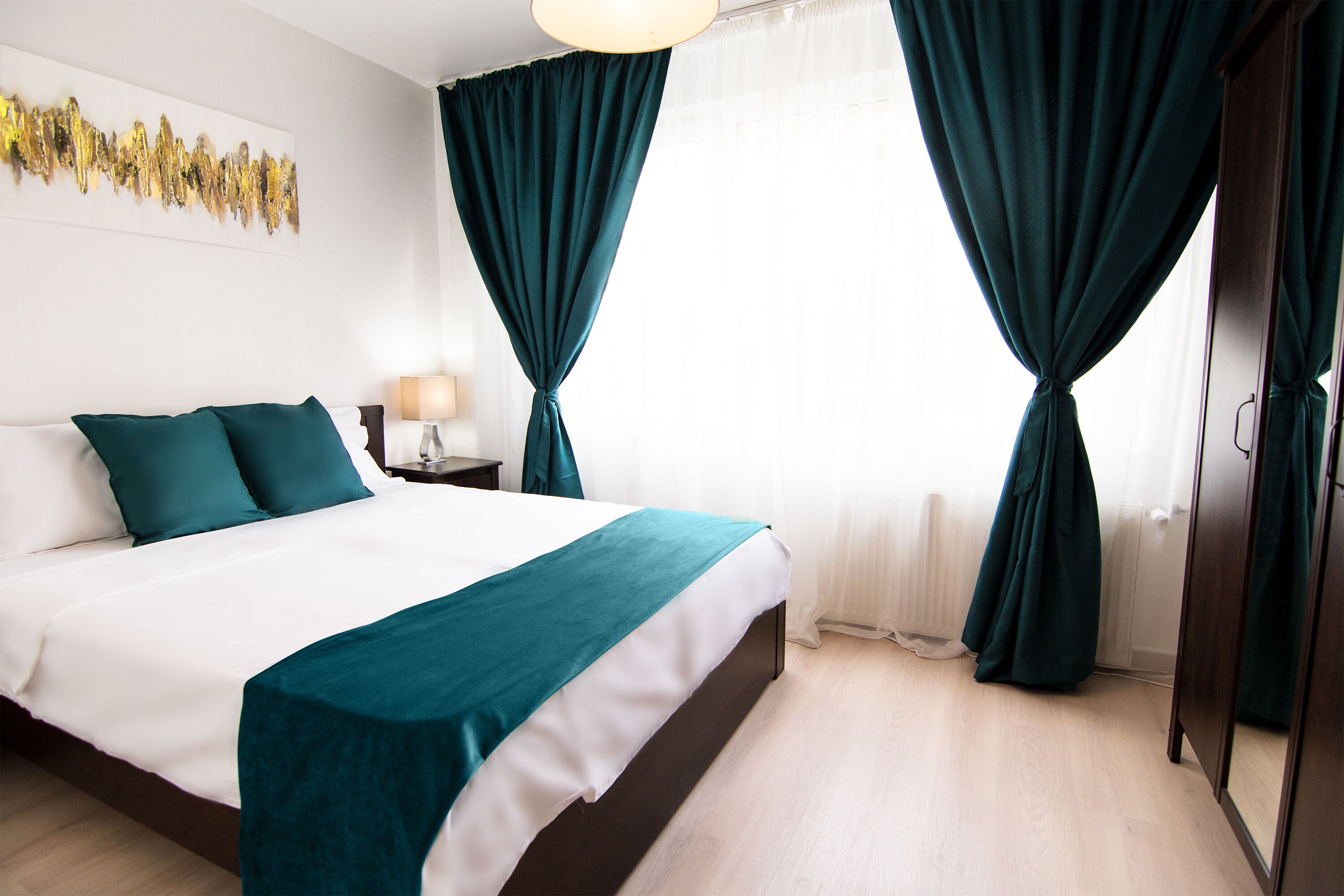 Habitación de hotel con una cama grande, cortinas verde azulado, suelo de madera y una pared blanca con una obra de arte abstracta dorada.