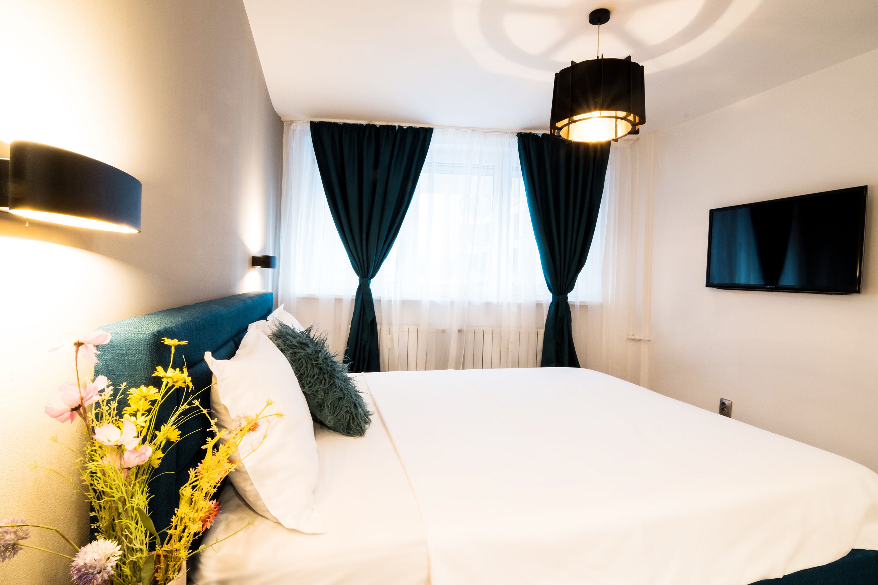 A modern hotel room with a large bed, dark curtains, a flat-screen TV, and a wall-mounted light fixture.