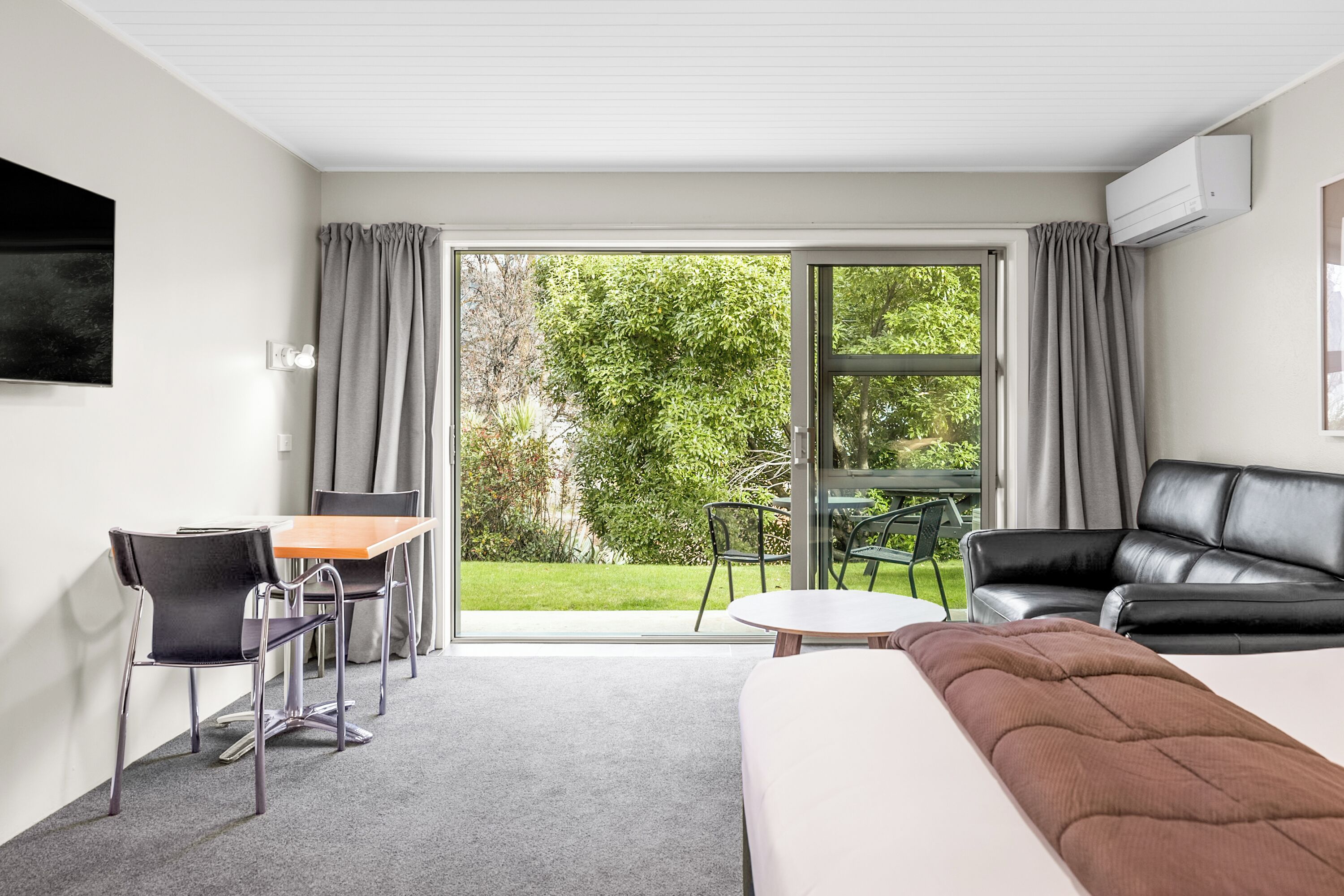 A hotel room with a bed, a desk with chairs, a sofa, and a large sliding glass door leading to a garden.