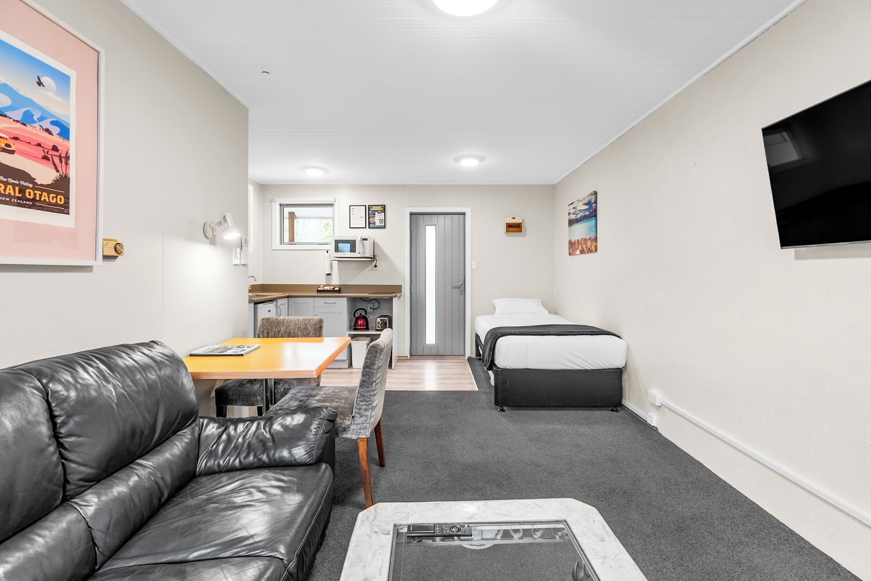 A compact room with a leather sofa, a small dining table, a bed, and a TV mounted on the wall.