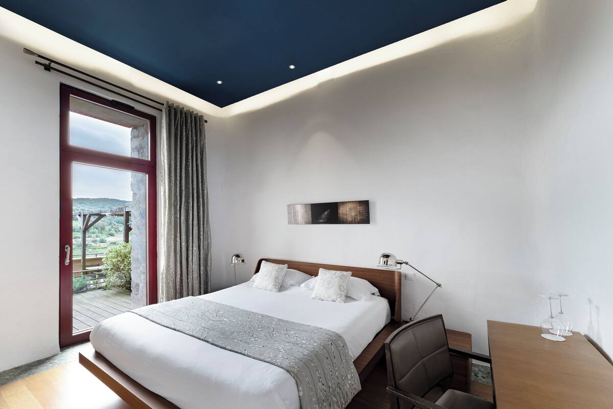 A modern bedroom with a large bed, a desk, and a view of the outdoors.