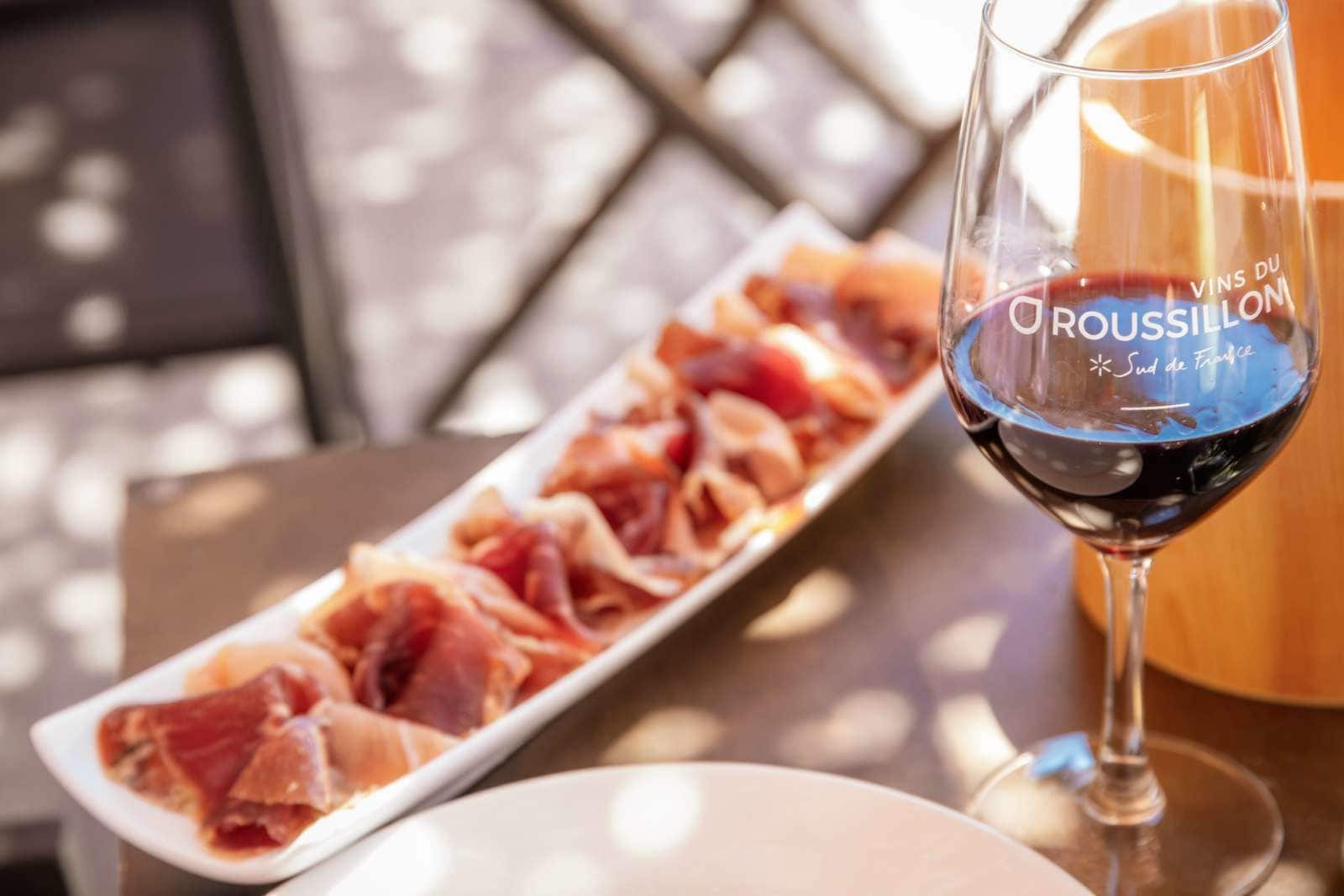 Ett glas rött vin med en etikett som visar texten ”Vins du Roussillon Sud de France”, placerat bredvid en vit, rektangulär tallrik med skivad charkuterier.