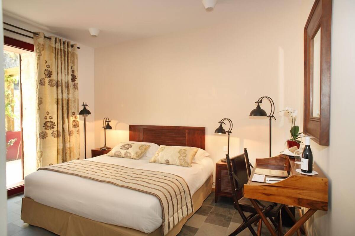 A bedroom with a large bed, two bedside tables with lamps, a wooden desk with a chair, and a window with curtains.