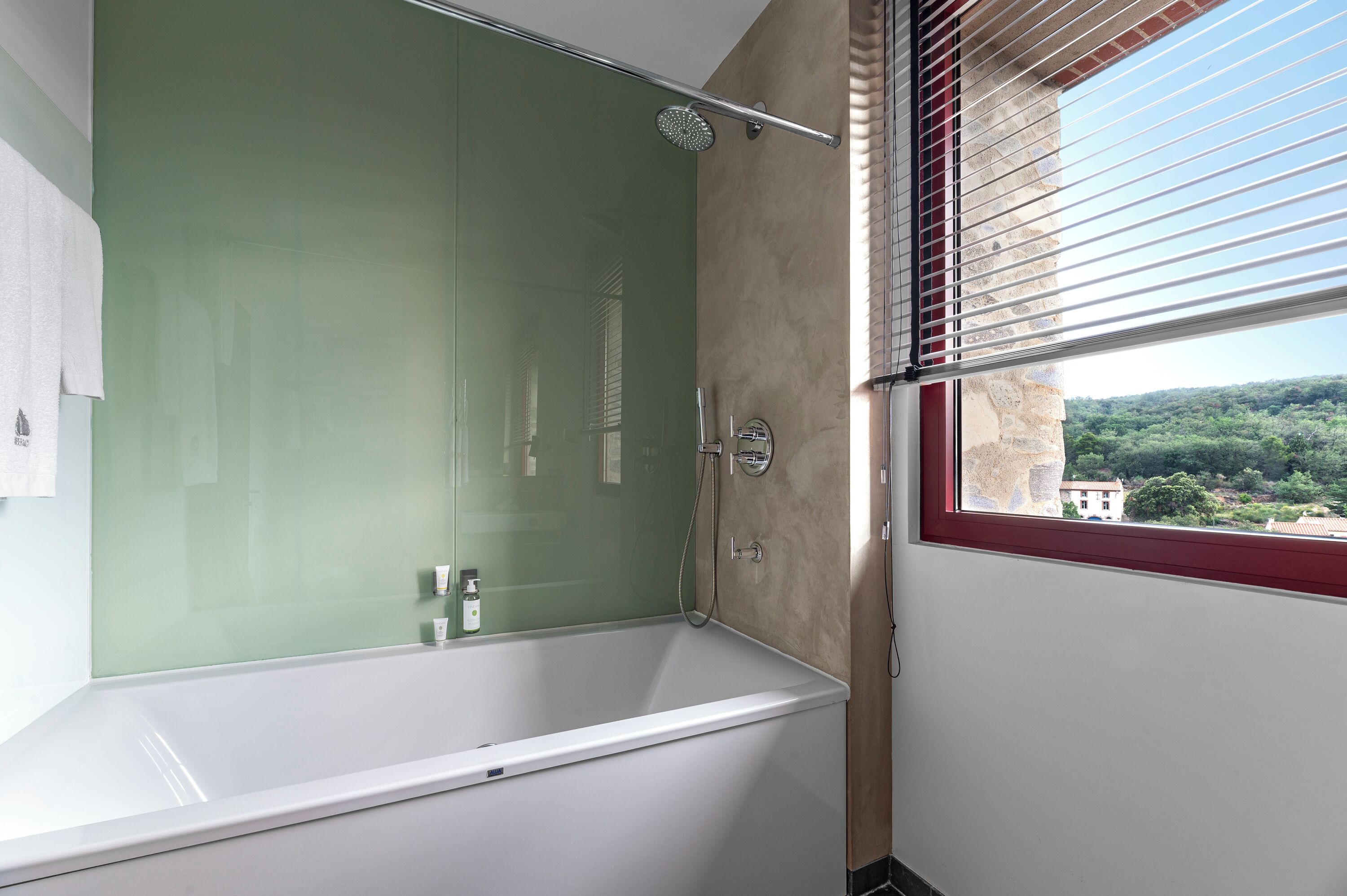 A modern bathroom with a glass-enclosed shower, a white bathtub, and a view of a green landscape through a window.