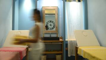 A room with a large wall clock, two sun loungers, a wooden shelf with bottles, and a mirror.