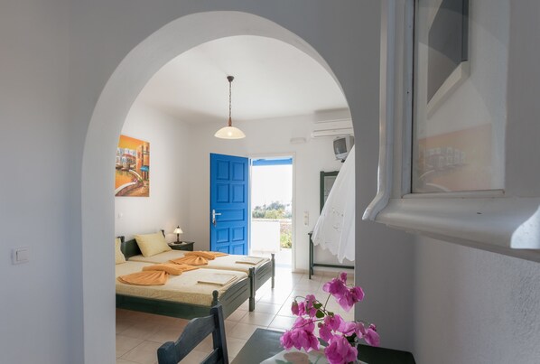 A room with a bed, a table with a vase of flowers, a blue door, and a view of the outside.