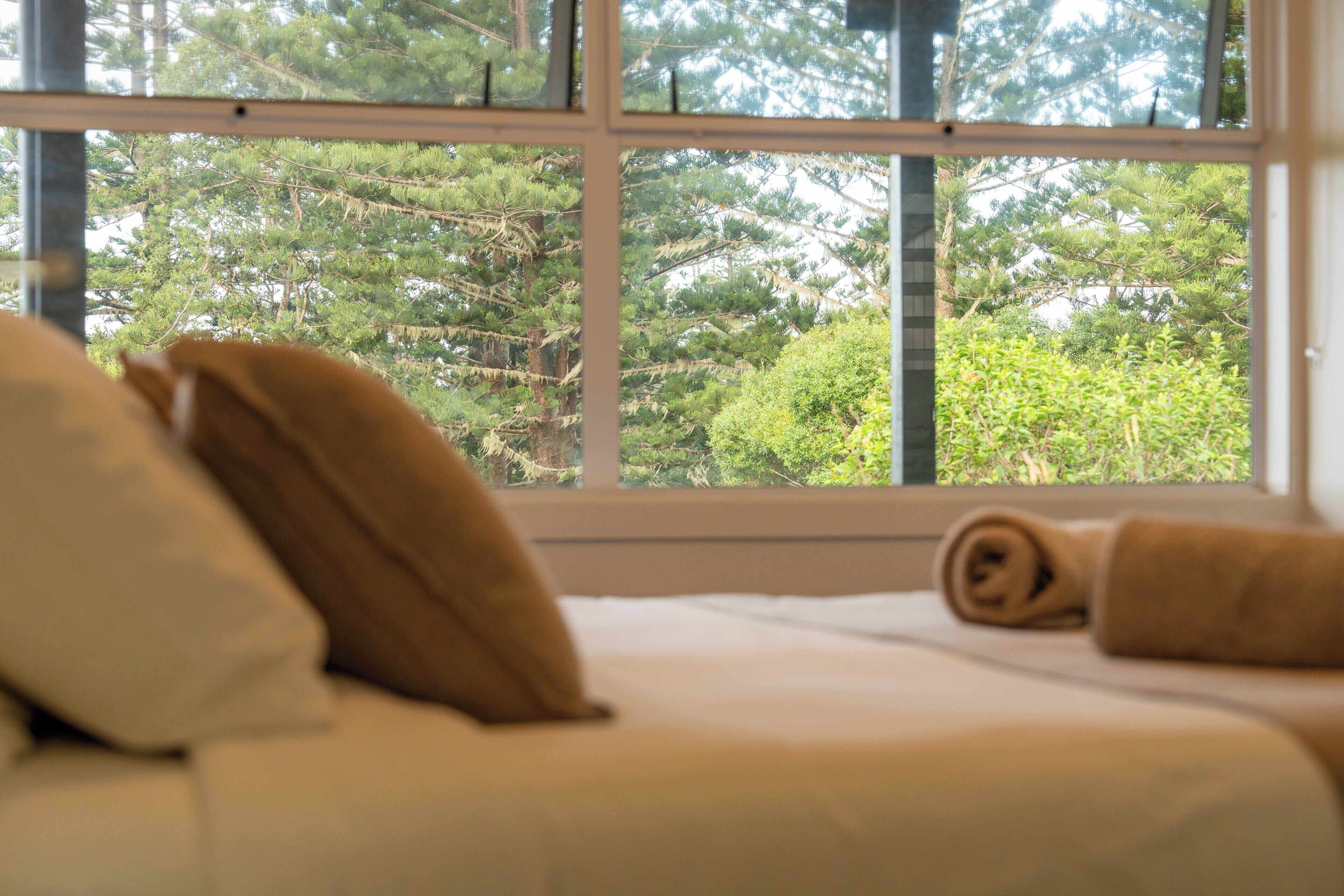 Uma cama com almofadas e uma toalha enrolada, uma janela com vista para árvores.