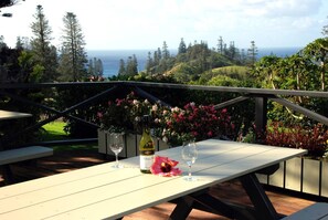 Une table avec une bouteille de vin et deux verres, placée devant un paysage pittoresque avec des collines et l’océan.