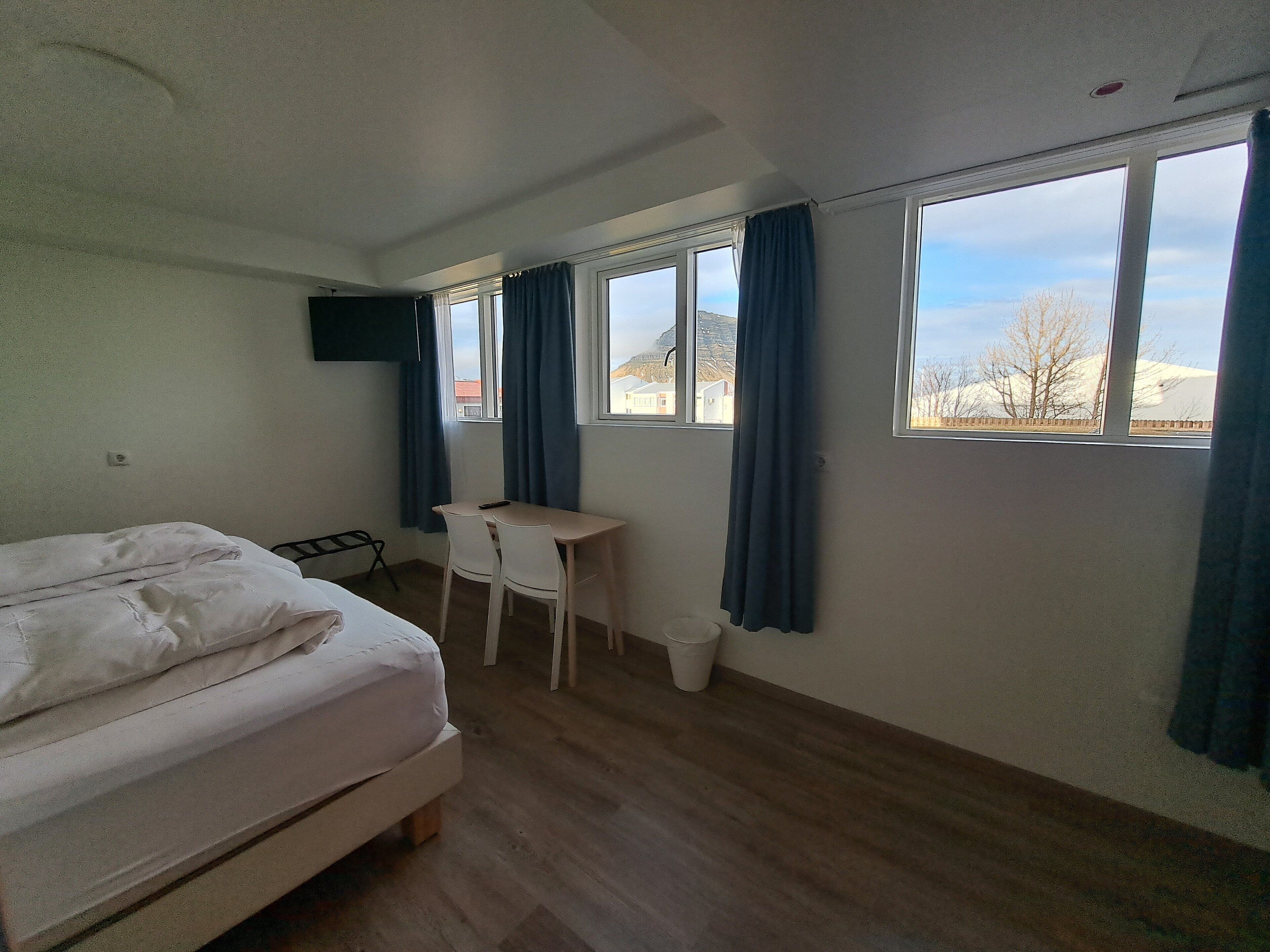 A bedroom with a bed, a desk, and two chairs. There are windows with curtains, a television mounted on the wall, and a view of a mountain and buildings outside.