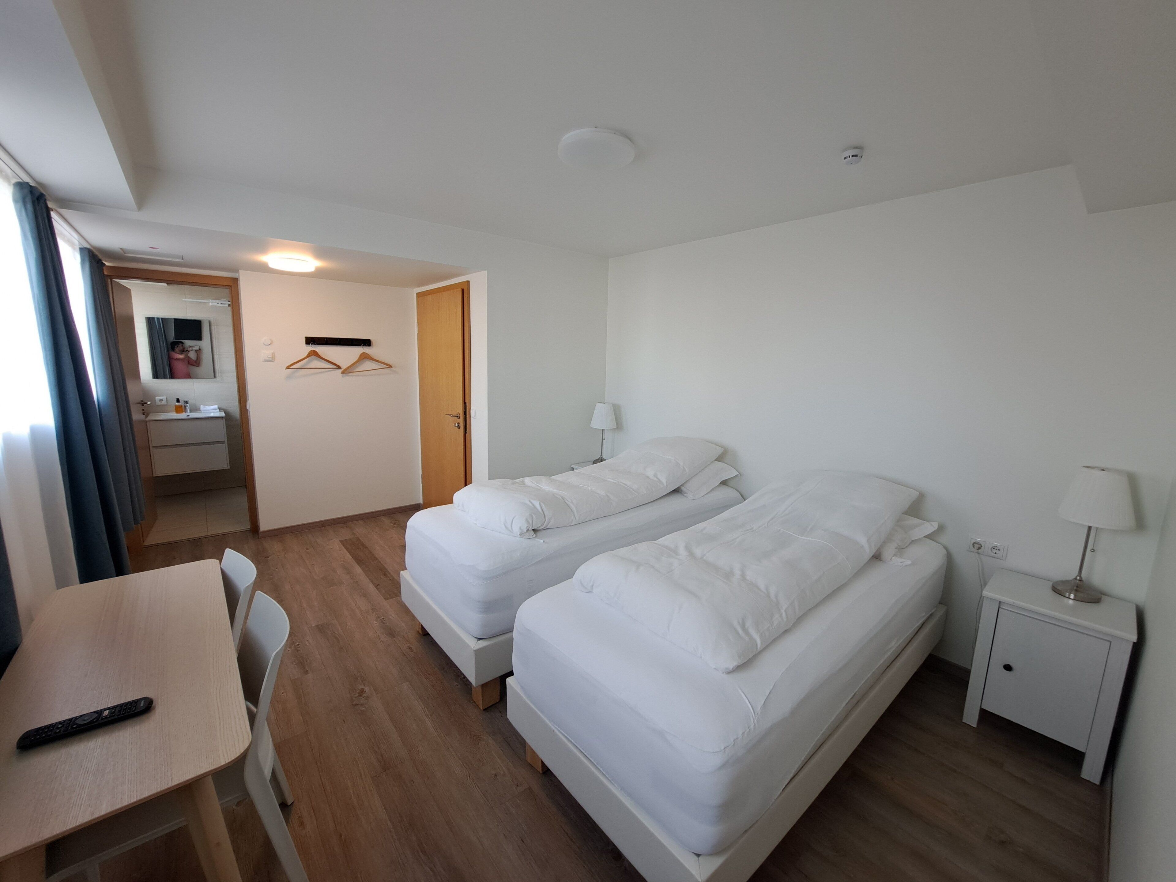 A hotel room with two beds, a desk, a chair, a bedside table with a lamp, and a bathroom visible through an open door.