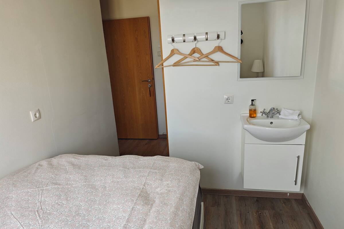 Un dormitorio con cama, un lavamanos blanco, un espejo y perchas de madera en la pared.